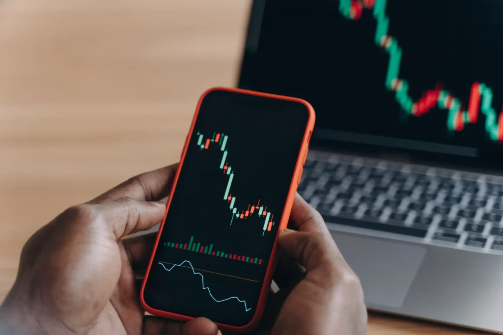 Close-up of African man’s hands holding a smartphone displaying stock market charts in front of a laptop.