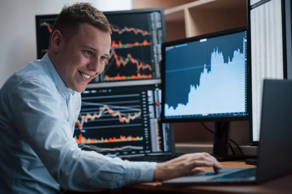 Businessman sitting in office with multiple monitors showing financial charts and stock market data.